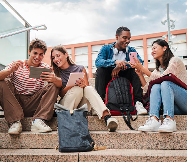Group of young friends sitting on steps looking at devices with their backpacks nearby