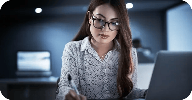 an image of a woman writing in a notebook while sitting in front of a laptop