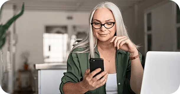 an image of a middle-aged woman sitting in front of a laptop while holding and browsing a mobile phone
