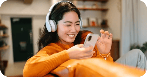 an image of a woman sitting on a couch wearing headphones and choosing music on her smartphone