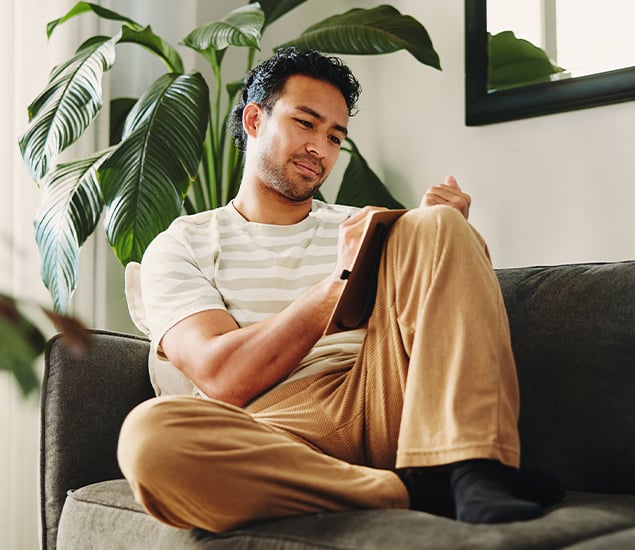 A man sitting on a couch in a bright living room, writing in a journal with a tablet on his lap, surrounded by green houseplants.